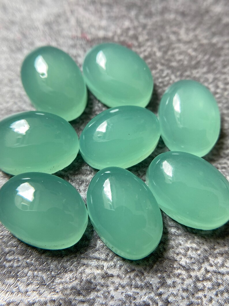 May include: Eight oval-shaped, polished gemstones in a soft, seafoam green hue. The stones have a smooth, glossy surface, reflecting light. The gemstones are arranged in a scattered pattern, showcasing their individual shapes and color.