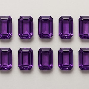 May include: Ten rectangular, faceted amethyst gemstones arranged in two rows of five. The gems have a deep purple hue and are cut in an emerald shape, showcasing their internal facets. The background is a plain, light gray.