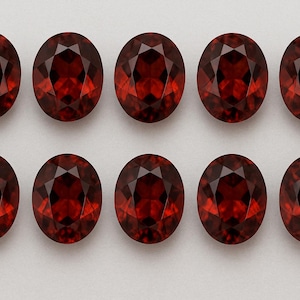 May include: Ten oval-cut, faceted, deep red gemstones are arranged in two rows of five. The gems have a brilliant, polished appearance, reflecting light. The stones are set against a neutral, light background.