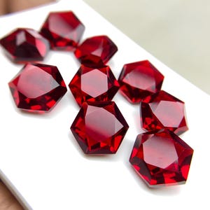 May include: Several faceted, hexagonal-shaped, deep red gemstones are displayed. The gems are arranged on a white surface, highlighting their brilliant cut and rich colour. The gemstones appear to be of high quality, with a polished finish.