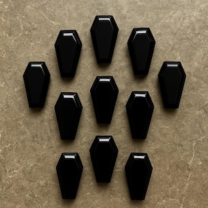 May include: Eleven small, glossy black coffin-shaped objects are arranged on a textured, neutral-toned surface. The coffins are evenly spaced and reflect light, highlighting their shape and design. The overall theme suggests a gothic or Halloween-related product.
