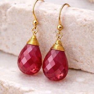 May include: Gold-coloured drop earrings featuring faceted, teardrop-shaped, red gemstones. The gemstones are wire-wrapped in gold. The earrings hang from gold ear wires. The earrings are displayed on a light-coloured, textured surface.