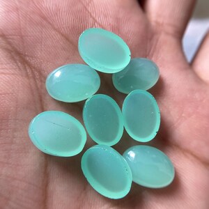 May include: A collection of eight oval-shaped, light teal gemstones. The polished stones are smooth and translucent, with a subtle sheen. The gemstones are held in a hand, showcasing their color and shape.