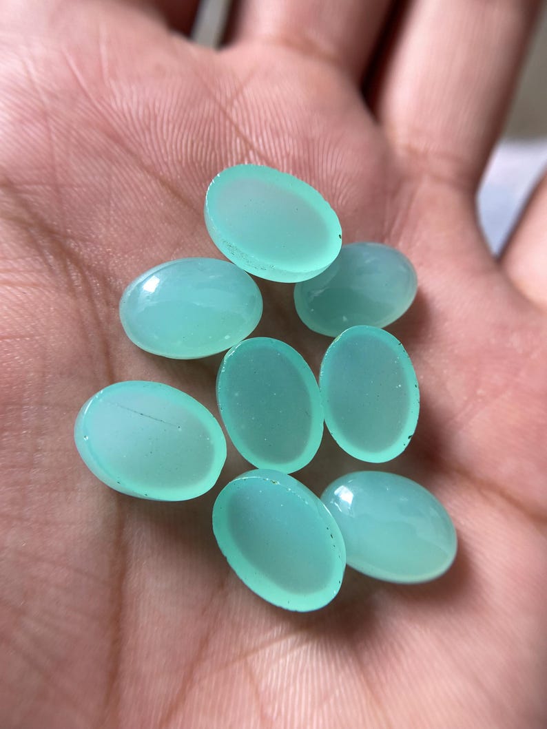 May include: A collection of eight oval-shaped, light teal gemstones. The polished stones are smooth and translucent, with a subtle sheen. The gemstones are held in a hand, showcasing their color and shape.