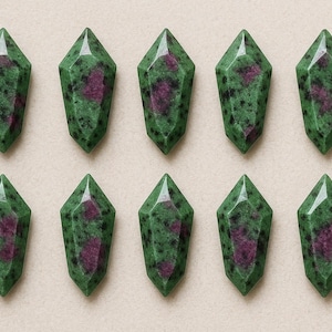 May include: Ten faceted, elongated gemstone crystals. Each crystal is a deep green with dark speckles and a central, irregular patch of deep pink. The crystals are arranged in two rows of five.