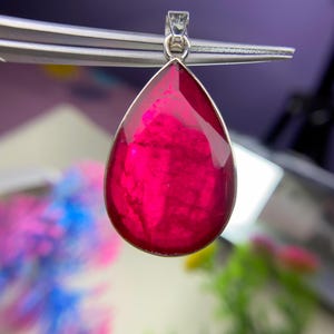 May include: A magenta teardrop pendant, held by tweezers. The gemstone is set in silver-coloured metal. The background is blurred, with hints of green and blue. The pendant's shape is reminiscent of a water droplet.