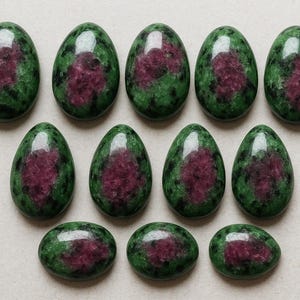 Peut inclure: Une collection de onze pierres précieuses ovales. Chaque pierre présente une base vert foncé avec une tache centrale irrégulière de rouge foncé. Les pierres sont disposées en trois rangées, les rangées supérieures et centrales étant ovales et la rangée inférieure plus arrondie.