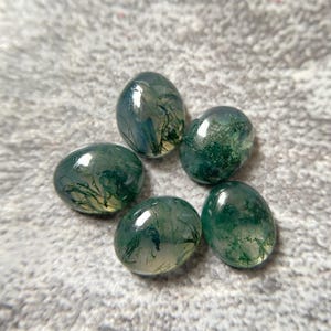 May include: Five polished, oval-shaped moss agate gemstones. The translucent stones display a mix of green, white, and blue inclusions, resembling miniature landscapes. The gemstones are arranged in a circular pattern.