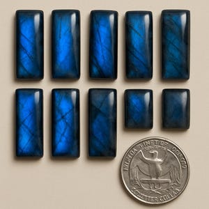 May include: A selection of rectangular labradorite gemstones displaying iridescent blue and teal colours. The stones are of varying sizes and shapes, with a coin for scale. The gemstones are arranged on a neutral background.