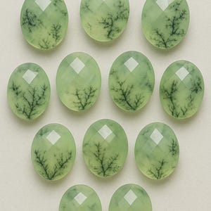 May include: A collection of eleven oval-shaped gemstones. Each gem is a pale green color with a faceted surface and a dark, tree-like inclusion pattern. The stones are arranged in a grid-like pattern.