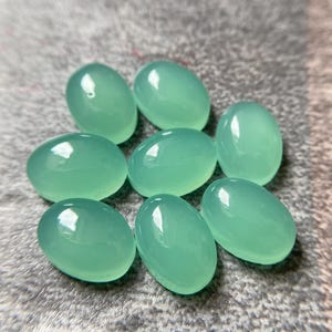 May include: Eight oval-shaped, polished gemstones in a soft, seafoam green hue. The stones are smooth and reflective, with a subtle sheen. They are arranged on a textured, gray surface.
