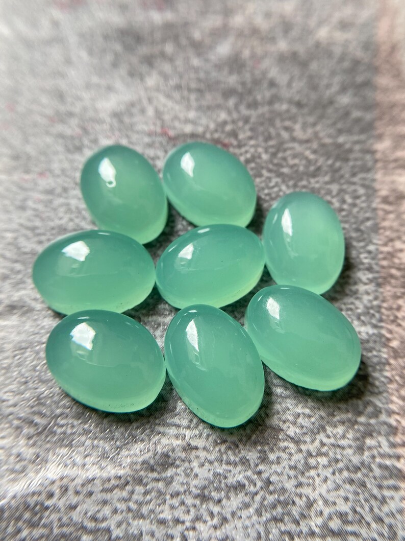May include: Eight oval-shaped, polished gemstones in a soft, seafoam green hue. The stones are smooth and reflective, with a subtle sheen. They are arranged on a textured, gray surface.