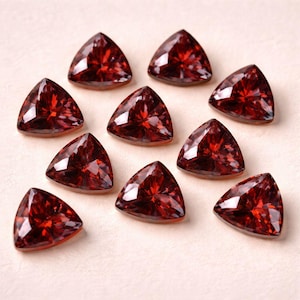 May include: Ten triangular, faceted, deep red gemstones are arranged on a light beige surface. The gems have a brilliant, reflective quality, with multiple facets catching the light. The overall arrangement suggests a collection of loose gemstones.