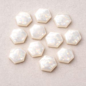 May include: A collection of ten iridescent, hexagonal-shaped beads. Each bead has a pearlescent, white surface with subtle light reflections. The beads are arranged on a neutral, textured fabric, suggesting a craft or jewelry-making theme.