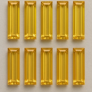 Puede incluir: Diez gemas rectangulares de color amarillo dorado están dispuestas en dos filas de cinco sobre un fondo neutro. Cada gema tiene una superficie facetada y pulida, que refleja la luz. Las gemas son de tamaño y forma similares, lo que sugiere una colección o un conjunto de componentes de joyería.