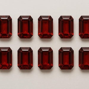 Könnte beinhalten: Zehn rechteckige, tiefrote Edelsteine mit Smaragdschliff sind in zwei Reihen zu je fünf angeordnet. Die Edelsteine haben eine satte, gesättigte Farbe und sind vor einem neutralen, hellen Hintergrund platziert. Die Facetten fangen das Licht ein und erzeugen einen funkelnden Effekt.