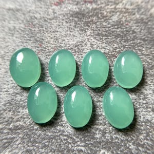May include: Seven oval-shaped, polished gemstones in a soft, seafoam green hue. The stones are smooth and reflective, with a subtle translucence. They are arranged on a textured, gray surface, showcasing their natural beauty and potential for jewelry making or crafting.