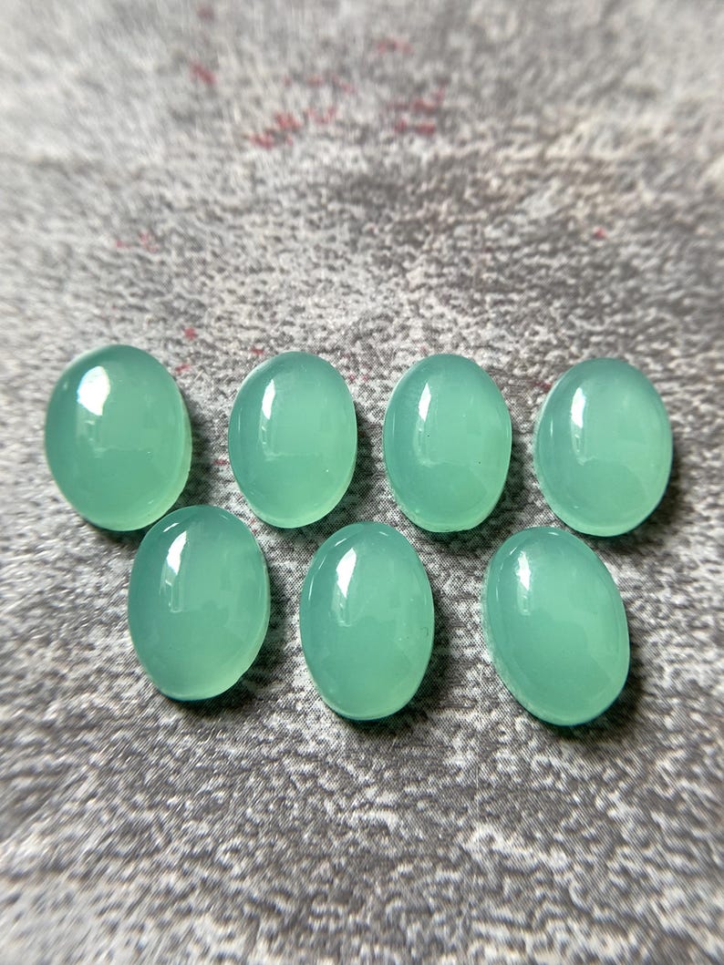 May include: Seven oval-shaped, polished gemstones in a soft, seafoam green hue. The stones are smooth and reflective, with a subtle translucence. They are arranged on a textured, gray surface, showcasing their natural beauty and potential for jewelry making or crafting.