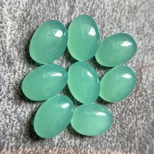 May include: Seven oval-shaped, polished gemstones in a soft, seafoam green hue. The stones are smooth and reflective, with a slight translucence. They are arranged in a circular pattern on a textured, gray surface.