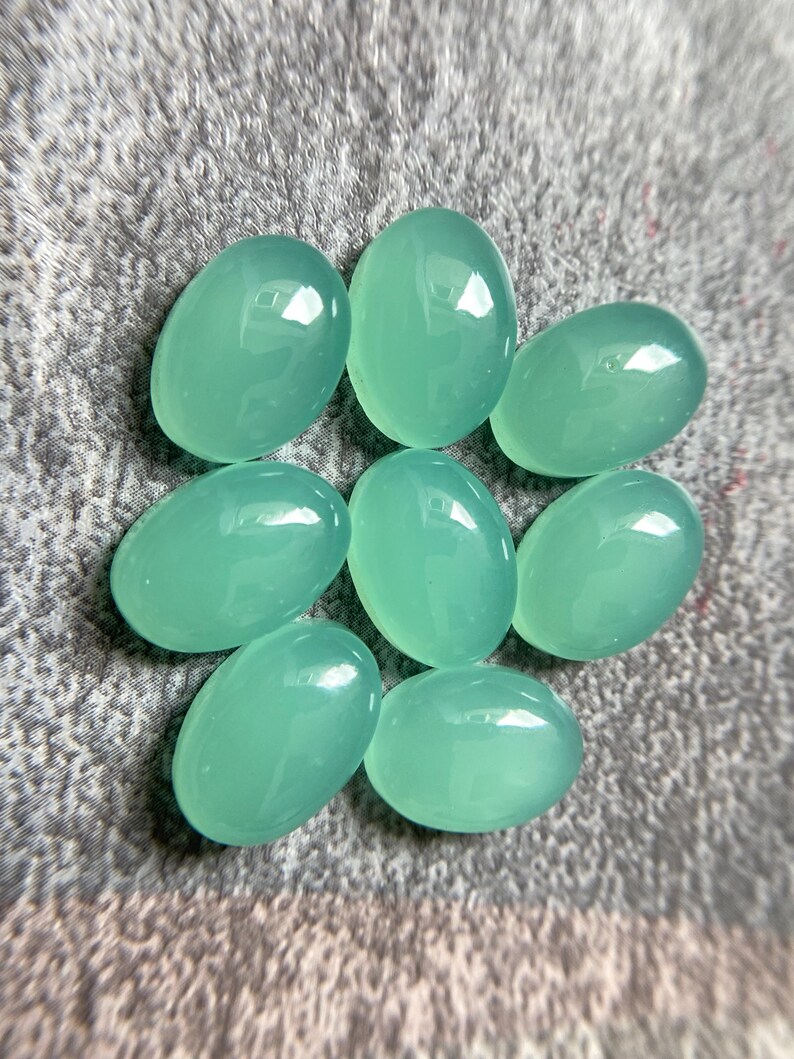 May include: Seven oval-shaped, polished gemstones in a soft, seafoam green hue. The stones are smooth and reflective, with a slight translucence. They are arranged in a circular pattern on a textured, gray surface.