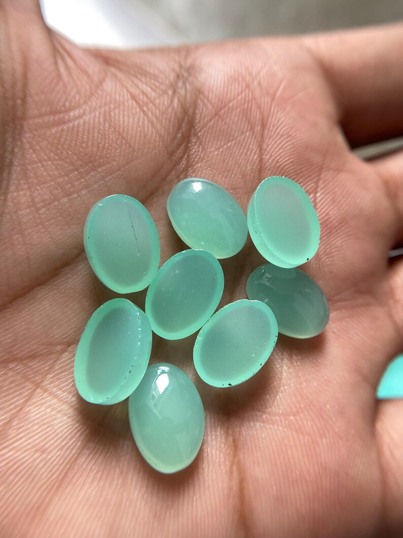 May include: A collection of eight oval-shaped, translucent, light-green gemstones. The stones have a smooth, polished surface and are displayed against a neutral background. These gemstones are ideal for jewelry making.