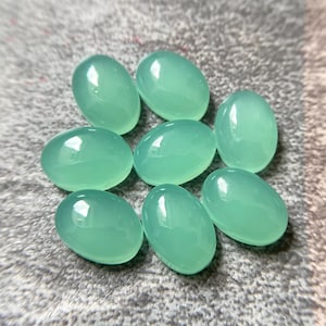 May include: Eight oval-shaped, polished gemstones in a light teal color. The stones are smooth and glossy, with a slightly translucent appearance. They are arranged on a textured, gray surface.