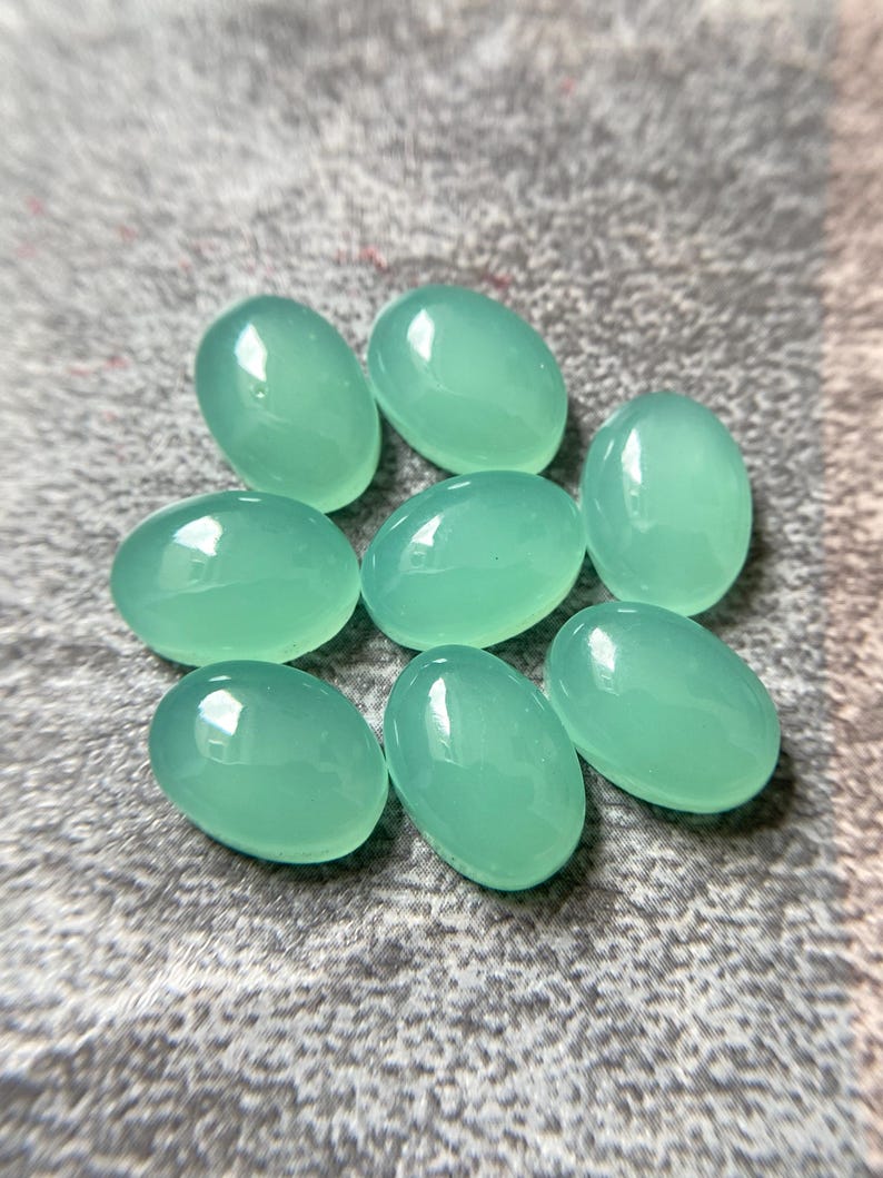 May include: Eight oval-shaped, polished gemstones in a light teal color. The stones are smooth and glossy, with a slightly translucent appearance. They are arranged on a textured, gray surface.