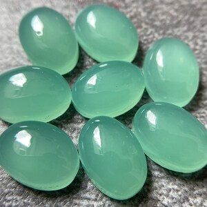 May include: Eight oval-shaped, polished gemstones in a soft, seafoam green hue. The stones have a smooth, glossy surface, reflecting light. The gemstones are arranged in a scattered pattern, showcasing their individual shapes and color.