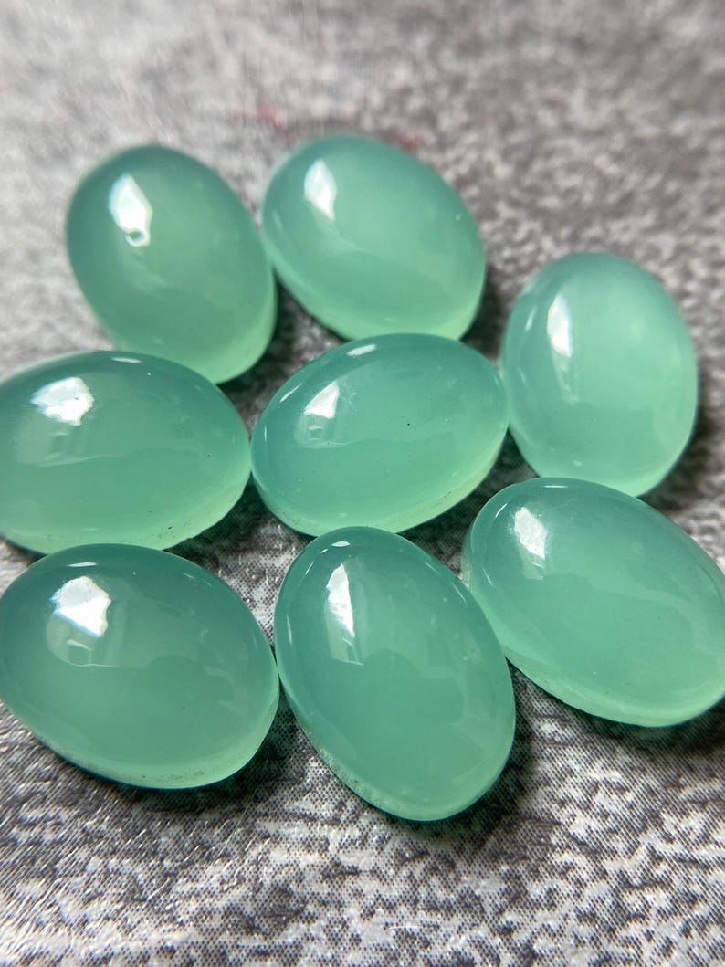May include: Eight oval-shaped, polished gemstones in a soft, seafoam green hue. The stones have a smooth, glossy surface, reflecting light. The gemstones are arranged in a scattered pattern, showcasing their individual shapes and color.
