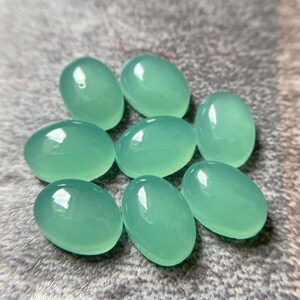 May include: Eight oval-shaped, polished gemstones in a soft, seafoam green hue. The stones are smooth and reflective, with a subtle sheen. They are arranged on a textured, gray surface.