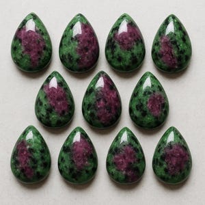 May include: Ten teardrop-shaped ruby zoisite gemstones. Each stone features a vibrant mix of deep green and speckled black with a central patch of rich, dark pink. The polished stones are arranged in a grid pattern.