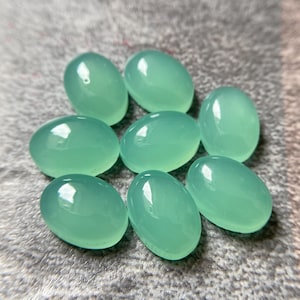 May include: Eight oval-shaped, polished gemstones in a soft, seafoam green hue. The stones have a smooth, glossy surface, reflecting light. The gemstones are arranged on a textured, gray surface. These stones are suitable for jewelry making.