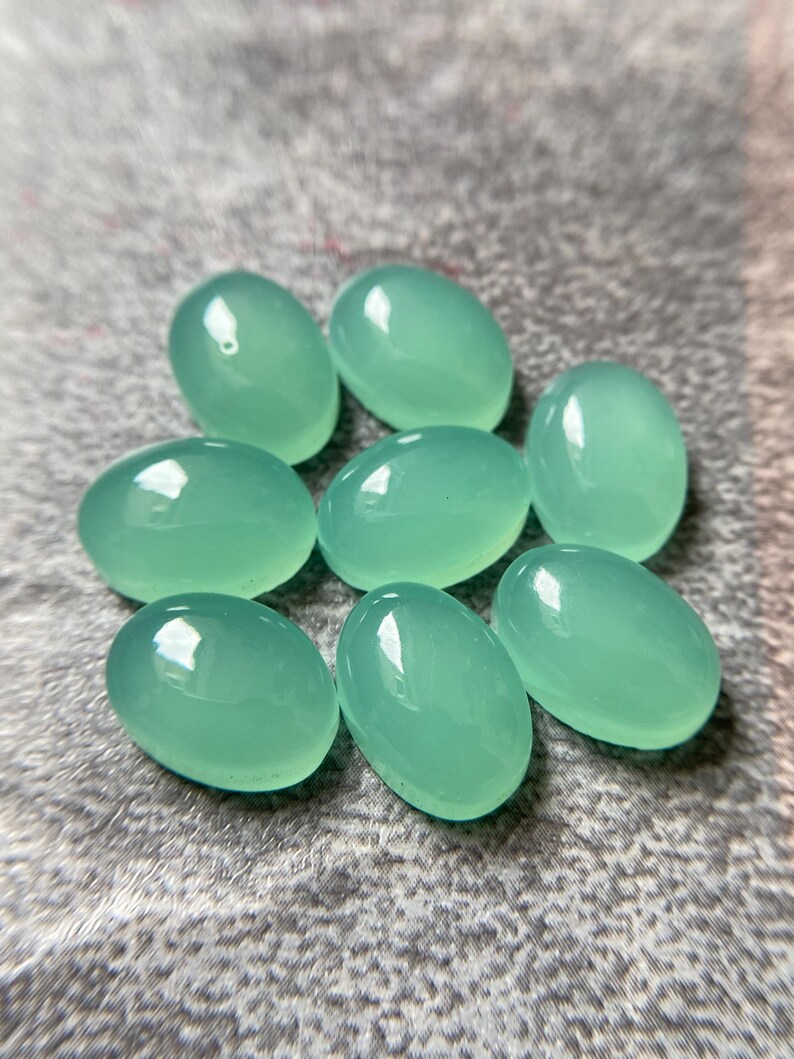 May include: Eight oval-shaped, polished gemstones in a soft, seafoam green hue. The stones have a smooth, glossy surface, reflecting light. The gemstones are arranged on a textured, gray surface. These stones are suitable for jewelry making.