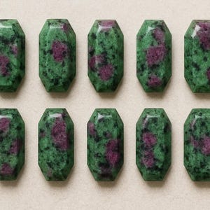 May include: Ten green and red ruby zoisite gemstone beads. Each bead is rectangular with faceted edges. The green stone has red inclusions, and the beads are arranged in two rows of five.