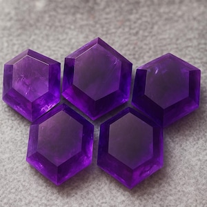 May include: Five faceted, hexagonal amethyst gemstones are arranged on a gray surface. The gemstones are a deep purple color, with a polished, reflective surface. The arrangement suggests a star or flower shape.
