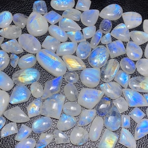 May include: A selection of polished moonstone gemstones in diverse forms, such as oval, teardrop, and rectangular. The stones exhibit a milky white base with iridescent blue and gold flashes. The gemstones are arranged on a dark surface.
