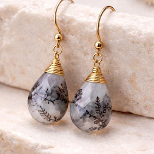 May include: A pair of gold-toned dangle earrings. Each earring features a teardrop-shaped, faceted stone with gray and black dendritic inclusions. The stones are capped with gold wire and suspended from gold-filled ear wires.