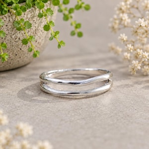 May include: A silver-toned ring featuring a double-band design with a smooth, polished finish. The ring consists of two parallel, slightly curved bands that are connected, creating an open space between the two distinct metal lines.