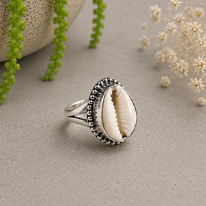 Könnte beinhalten: Ein Silberring mit einer zentralen weißen Kaurimuschel. Die Muschel ist in eine perlenartige Silberfassung mit polierter Oberfläche eingefasst. Der Ringband ist glatt und silbern und bildet einen klaren Kontrast zum strukturierten, organischen Aussehen der Muschel.