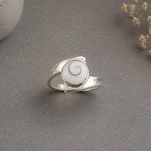 May include: A silver ring featuring a circular white shiva eye shell inlay. The shell displays a natural brown spiral pattern centred on a smooth, polished surface. The metal band has a wide, open-ended design that tapers towards the setting.