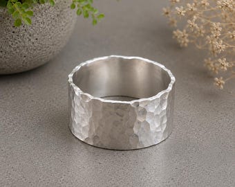 May include: A wide silver ring featuring a textured, hammered finish across its entire outer surface. The band has a consistent width and a polished, reflective metallic appearance, showcasing a series of small, irregular indentations.