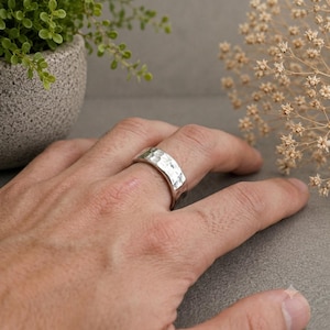 May include: A silver ring with a textured, hammered finish worn on a finger. The band has a wide, flat profile with irregular indentations across the surface, creating a reflective, metallic appearance that catches the light.