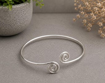 May include: A silver-toned metal cuff bracelet featuring a smooth, circular band that terminates at both ends in a decorative, tightly coiled spiral design. The bracelet has an open-ended construction, allowing for adjustment to fit around the wrist.