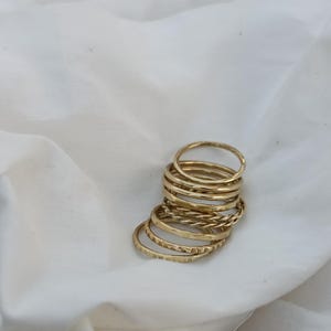 Puede incluir: Una pila de anillos dorados con diversas texturas, incluyendo diseños lisos, martillados y trenzados. Los anillos se exhiben sobre un fondo de tela blanca, creando una presentación simple y elegante.