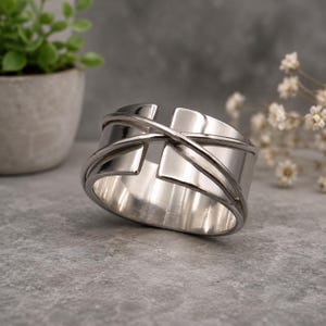 Puede incluir: Una pulsera tipo brazalete ancha de tono plateado pulido con un diseño de banda abierta. La superficie está acentuada con dos hilos finos superpuestos que se envuelven diagonalmente por la parte frontal, creando un aspecto dimensional.