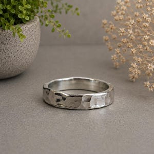May include: A silver-toned ring featuring a distinct hammered texture across its outer surface. The band has a rounded, irregular profile with a polished metallic finish that reflects light across its uneven, indented facets and smooth interior.