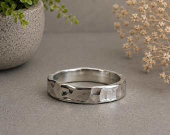 May include: A silver-toned ring featuring a distinct hammered texture across its outer surface. The band has a rounded, irregular profile with a polished metallic finish that reflects light across its uneven, indented facets and smooth interior.