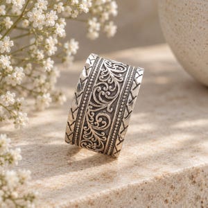 Puede incluir: Un anillo ancho de plata con un intrincado patrón de volutas en relieve en el centro, flanqueado por bordes de cuentas y bandas exteriores con un diseño grabado en diagonal. El metal tiene un acabado oscuro y oxidado en las zonas.