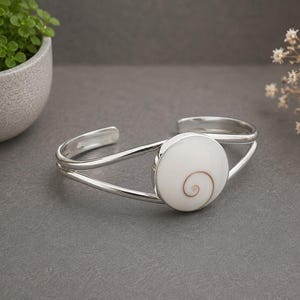 Puede incluir: Una pulsera de plata tipo brazalete con una pieza central circular de concha shiva blanca. La concha tiene un patrón de espiral marrón natural en el centro. La banda de la pulsera está hecha de dos hilos de plata finos y pulidos.