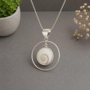 Puede incluir: Un collar de plata con un colgante circular que sostiene una concha Shiva redonda y blanca con un patrón de espiral marrón natural. El colgante cuelga de una delicada cadena de plata, creando una pieza de joyería geométrica y equilibrada.